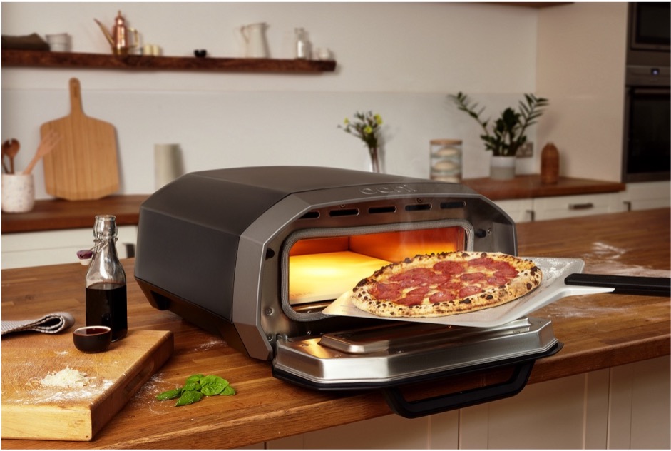 Her ser du en elektrisk pizza ovn fra Ooni der er placeret i et køkken. der er en pizza på vej ind i ovnen ved hjælp af en pizzaspade