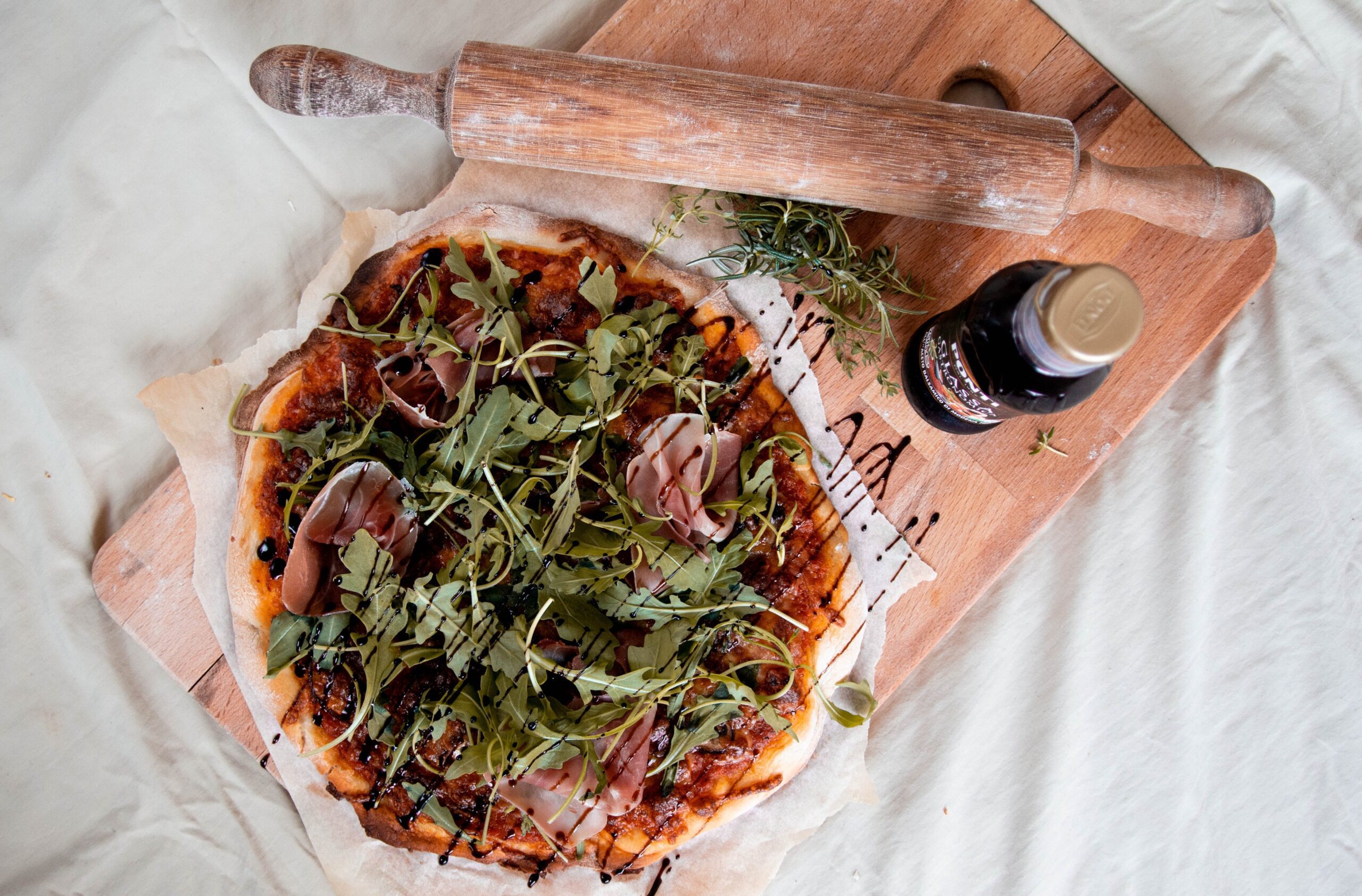 På billedet ses en Prosciutto Pizza liggende på et skærrebræt. Ved siden af står en balsamico dressing.